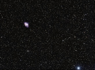 A vast expanse of space with numerous stars scattered across the dark sky. A nebula appears prominently on the upper left side, glowing with hues of purple and blue.