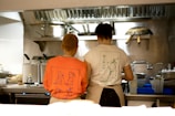 Two people are standing in a professional kitchen, facing away from the camera. They wear t-shirts with artistic designs on the back, one in orange and the other in white. Various kitchen utensils and equipment, such as pots and ladles, are visible around them.