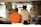 Two people are standing in a professional kitchen, facing away from the camera. They wear t-shirts with artistic designs on the back, one in orange and the other in white. Various kitchen utensils and equipment, such as pots and ladles, are visible around them.