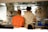 Two people are standing in a professional kitchen, facing away from the camera. They wear t-shirts with artistic designs on the back, one in orange and the other in white. Various kitchen utensils and equipment, such as pots and ladles, are visible around them.