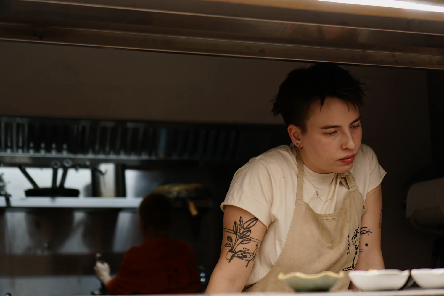 A young person with a tattooed forearm standing in a kitchen