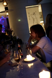 A dimly lit bar with a few people seated at a counter. Candles on the bar top create a warm, intimate atmosphere. A woman in a white shirt is smiling and engaged in conversation, holding a drink. Various drinks, including a beer bottle and wine glasses, are placed on the counter. Hanging light fixtures add to the ambient lighting.