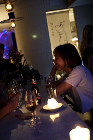 A dimly lit bar with a few people seated at a counter. Candles on the bar top create a warm, intimate atmosphere. A woman in a white shirt is smiling and engaged in conversation, holding a drink. Various drinks, including a beer bottle and wine glasses, are placed on the counter. Hanging light fixtures add to the ambient lighting.