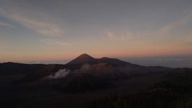 A picturesque landscape featuring a majestic mountain with a conical shape in the background. The foreground includes another rugged volcanic structure with a visible plume of smoke or steam rising from it. The sky is a beautiful gradient of colors transitioning from pink and orange near the horizon to a deeper blue as it ascends.