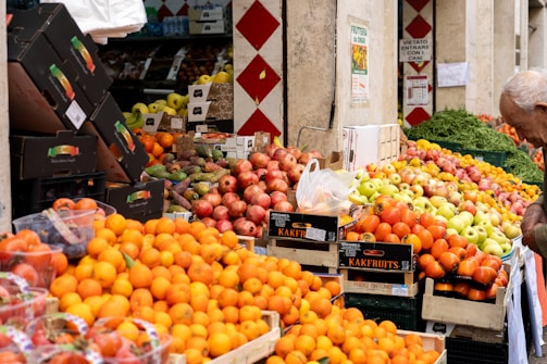A vibrant outdoor fruit market featuring piles of fresh produce including oranges, apples, and pomegranates. Wooden crates and cardboard boxes are used for displaying the fruits, and a variety of colors can be seen amongst the stacked goods. A man on the right is inspecting or selecting items. The market stalls are partially shaded by a building and surrounded by more crates of produce.