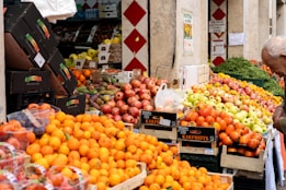 A vibrant outdoor fruit market featuring piles of fresh produce including oranges, apples, and pomegranates. Wooden crates and cardboard boxes are used for displaying the fruits, and a variety of colors can be seen amongst the stacked goods. A man on the right is inspecting or selecting items. The market stalls are partially shaded by a building and surrounded by more crates of produce.