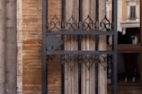 A detailed view of an ornate, black wrought iron gate installed against a textured brick and stone wall. The design of the gate features elegant, symmetrical patterns with curved motifs. Behind the gate, there's a reflection of a building facade with columns and windows, visible in the glass section.