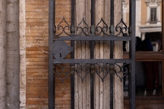 A detailed view of an ornate, black wrought iron gate installed against a textured brick and stone wall. The design of the gate features elegant, symmetrical patterns with curved motifs. Behind the gate, there's a reflection of a building facade with columns and windows, visible in the glass section.