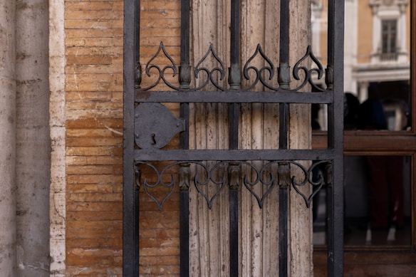 A detailed view of an ornate, black wrought iron gate installed against a textured brick and stone wall. The design of the gate features elegant, symmetrical patterns with curved motifs. Behind the gate, there's a reflection of a building facade with columns and windows, visible in the glass section.