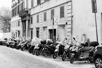 A row of parked scooters lines a cobblestone street adjacent to an old building with shuttered windows and balconies. A parking meter sign is visible, indicating ticket requirements. A few cars are also parked along the street.