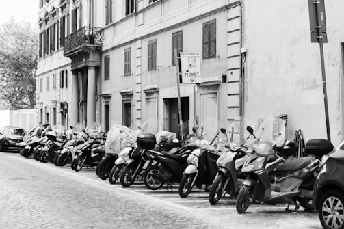 A row of parked scooters lines a cobblestone street adjacent to an old building with shuttered windows and balconies. A parking meter sign is visible, indicating ticket requirements. A few cars are also parked along the street.