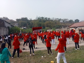 A large group of people is participating in an outdoor exercise session. Most are wearing red shirts and some have head coverings. They appear to be in a rural area, surrounded by greenery and small structures, with a stage in the background.