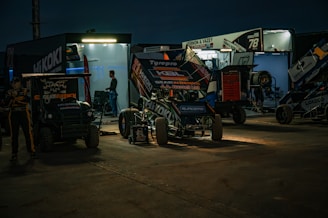 A nighttime scene with a racing pit stop. Several racing vehicles, including sprint cars, are parked in front of well-lit trailers. Mechanics and team members are seen working in the background. The area is illuminated by artificial lights, casting dramatic shadows on the ground.
