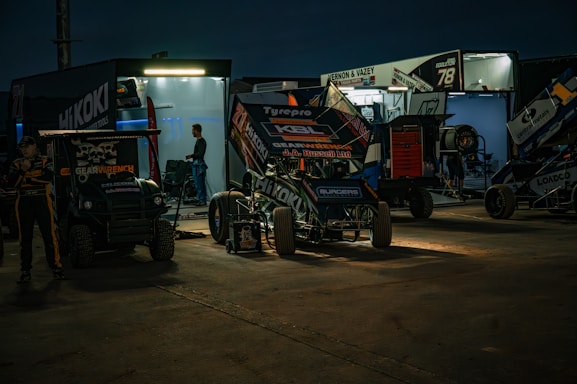 A nighttime scene with a racing pit stop. Several racing vehicles, including sprint cars, are parked in front of well-lit trailers. Mechanics and team members are seen working in the background. The area is illuminated by artificial lights, casting dramatic shadows on the ground.