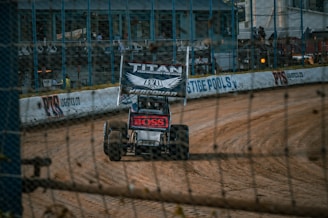 A dirt track race car speeds along a sandy pathway within an enclosed racing circuit. The car features sponsors’ logos and is surrounded by a barrier with advertisements displayed on it. Spectators can be seen in the background behind the fence.
