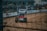 A dirt track race car speeds along a sandy pathway within an enclosed racing circuit. The car features sponsors’ logos and is surrounded by a barrier with advertisements displayed on it. Spectators can be seen in the background behind the fence.