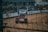 A dirt track race car speeds along a sandy pathway within an enclosed racing circuit. The car features sponsors’ logos and is surrounded by a barrier with advertisements displayed on it. Spectators can be seen in the background behind the fence.