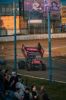 A race car with prominent advertising decals is driving on a dirt track. A crowd of spectators watches from behind a wire fence. The scene is illuminated by large screens showing more racing footage and a sign advertising an event called 'The Summer of More'. Shipping containers are visible in the background.