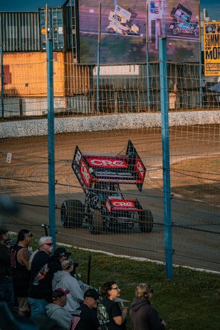 A race car with prominent advertising decals is driving on a dirt track. A crowd of spectators watches from behind a wire fence. The scene is illuminated by large screens showing more racing footage and a sign advertising an event called 'The Summer of More'. Shipping containers are visible in the background.