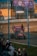 A race car with prominent advertising decals is driving on a dirt track. A crowd of spectators watches from behind a wire fence. The scene is illuminated by large screens showing more racing footage and a sign advertising an event called 'The Summer of More'. Shipping containers are visible in the background.