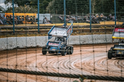 Two sprint cars are racing on a dirt track, with one car in blue and the other showcasing red accents. Spectators are sitting on lawn chairs behind a wire fence, watching the race. The track has visible tire marks and borders with a white barrier.