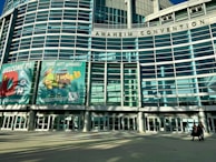 The image shows the facade of the Anaheim Convention Center with large glass panels and a modern architectural design. Two large banners are displayed, advertising an event called 'DCon' featuring toys, art, and apparel, with vibrant graphics of toys and collectibles. In the foreground, two people walk across a spacious plaza in front of the building.