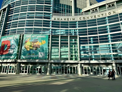 The image shows the facade of the Anaheim Convention Center with large glass panels and a modern architectural design. Two large banners are displayed, advertising an event called 'DCon' featuring toys, art, and apparel, with vibrant graphics of toys and collectibles. In the foreground, two people walk across a spacious plaza in front of the building.