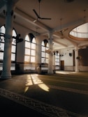 The image depicts a serene and spacious interior of a mosque with high ceilings and large arched windows casting patterned light onto the floor. The architecture is elegant with decorative columns and intricate lattice work along the walls. A person sits quietly against a column, bathed in the soft light from the windows.