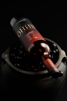 A wine bottle is positioned on a bed of dark grapes inside a rustic container. The bottle has a deep red label with gold lettering, and the overall setting is dimly lit, highlighting the glossy surface of the bottle and the grapes.