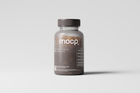A transparent brown plastic pill bottle with a dark cap is placed against a light gray background. The label on the bottle reads 'mocp' and includes additional text and a website link at the bottom. The bottle's contents appear to be small brown pills.