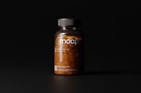 A dark-toned image showcasing a clear plastic bottle filled with orange pills under soft lighting. The bottle has a black cap and features minimalist labeling with the letters 'mocp' prominently displayed.