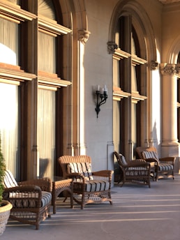 Several wicker chairs with striped cushions are arranged on a stone patio, basking in the warm glow of the afternoon sun. The patio is lined with large arched windows and stone columns, creating a serene and elegant outdoor setting.