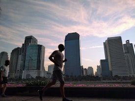 A silhouette of a person jogging in the foreground with a backdrop of tall modern skyscrapers. The sky is filled with soft clouds illuminated by a setting or rising sun casting a gentle light over the scene. Another person stands by observing in the background.