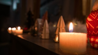 Candles are lit and placed on a surface, with a soft, warm glow illuminating the area. The background includes various decorative objects, including cone-shaped ornaments and a red and white patterned item, possibly fabric or knitwear.