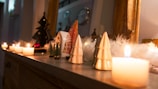 A cozy, festive scene with lit candles placed on a ledge, creating a warm glow. Small decorative objects, including ceramic Christmas trees and miniature pine trees, are arranged alongside fluffy white garland. The background includes more decor elements and mirrors, enhancing the holiday ambiance.