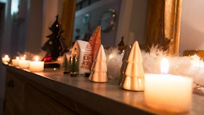 A cozy, festive scene with lit candles placed on a ledge, creating a warm glow. Small decorative objects, including ceramic Christmas trees and miniature pine trees, are arranged alongside fluffy white garland. The background includes more decor elements and mirrors, enhancing the holiday ambiance.