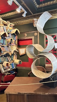 An eclectic library setting with a vintage typewriter, from which an oversized, spiraling paper emerges. The paper creates an artistic flow above a wooden desk. The bookshelf in the background is creatively chaotic, with books stacked in unusual angles, some open and fanned out. The room is adorned with red and dark green walls, gold framing, and spotlights adding a dramatic flair.