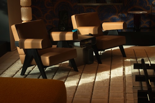 A cozy, stylish interior setting features two modern armchairs with tan leather upholstery and black legs. They are arranged on a cream, textured rug, creating a warm ambiance. The backdrop includes a dark, patterned wallpaper and a sleek, black table with a small lamp. Soft lighting casts shadows, enhancing the inviting atmosphere.