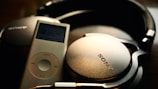 A pair of black Sony headphones is resting next to a silver iPod. The headphones have a cushioned headband and large ear cups, with the Sony logo prominently displayed. The iPod has a screen displaying music with a circular control pad below it.