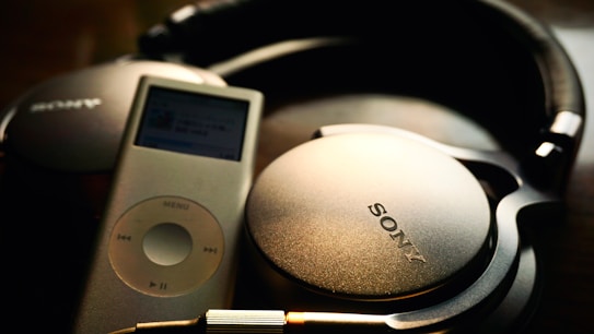 A pair of black Sony headphones is resting next to a silver iPod. The headphones have a cushioned headband and large ear cups, with the Sony logo prominently displayed. The iPod has a screen displaying music with a circular control pad below it.