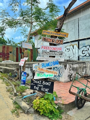 A collection of colorful wooden signs with handwritten text are affixed to a tree and a post in front of a building. The signs point in various directions and advertise different bars and services such as Stones Dorm Room, Stones Bar, Freedom Bar, Ibiza Pool Party, Blanco Bar, and Taxi Boat rentals. The area is adorned with graffiti, and there are green plants growing at the base of the post. The sky is partly cloudy, and the surroundings have a somewhat rustic and urban feel.