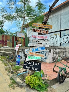 A collection of colorful wooden signs with handwritten text are affixed to a tree and a post in front of a building. The signs point in various directions and advertise different bars and services such as Stones Dorm Room, Stones Bar, Freedom Bar, Ibiza Pool Party, Blanco Bar, and Taxi Boat rentals. The area is adorned with graffiti, and there are green plants growing at the base of the post. The sky is partly cloudy, and the surroundings have a somewhat rustic and urban feel.