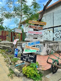 A collection of colorful wooden signs with handwritten text are affixed to a tree and a post in front of a building. The signs point in various directions and advertise different bars and services such as Stones Dorm Room, Stones Bar, Freedom Bar, Ibiza Pool Party, Blanco Bar, and Taxi Boat rentals. The area is adorned with graffiti, and there are green plants growing at the base of the post. The sky is partly cloudy, and the surroundings have a somewhat rustic and urban feel.