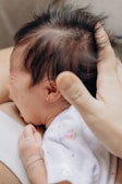 A newborn baby with dark hair is being gently held and comforted. The baby's small hand is visible, and they appear to be crying or fussing. The person holding the baby has their hand on the baby's head, providing a sense of care. The baby is wearing a white outfit with a floral pattern.