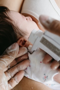 A close-up view of an infant having its ear pierced. The baby is resting on a white cloth with small floral designs. A person wearing gloves holds a piercing device against the infant's ear.