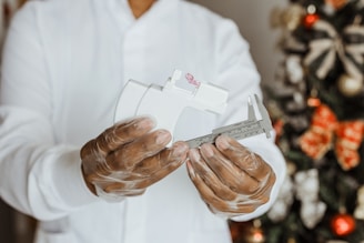 A person wearing a white lab coat and transparent gloves is holding a white and silver caliper with precise measurements. In the background, a decorated Christmas tree with red and gold ornaments is visible.