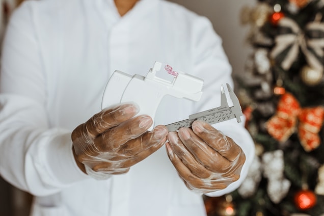 A person wearing a white lab coat and transparent gloves is holding a white and silver caliper with precise measurements. In the background, a decorated Christmas tree with red and gold ornaments is visible.