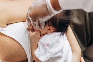 A baby is breastfeeding, held in an adult's arms. The adult is wearing protective gloves, indicating hygiene or medical care. The baby is dressed in floral-patterned clothing.