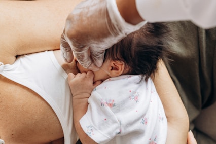 A baby is breastfeeding, held in an adult's arms. The adult is wearing protective gloves, indicating hygiene or medical care. The baby is dressed in floral-patterned clothing.