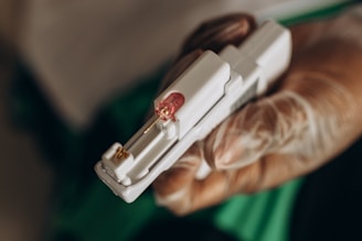A hand wearing a latex glove is holding a medical device with a small needle and a pink-colored cap.